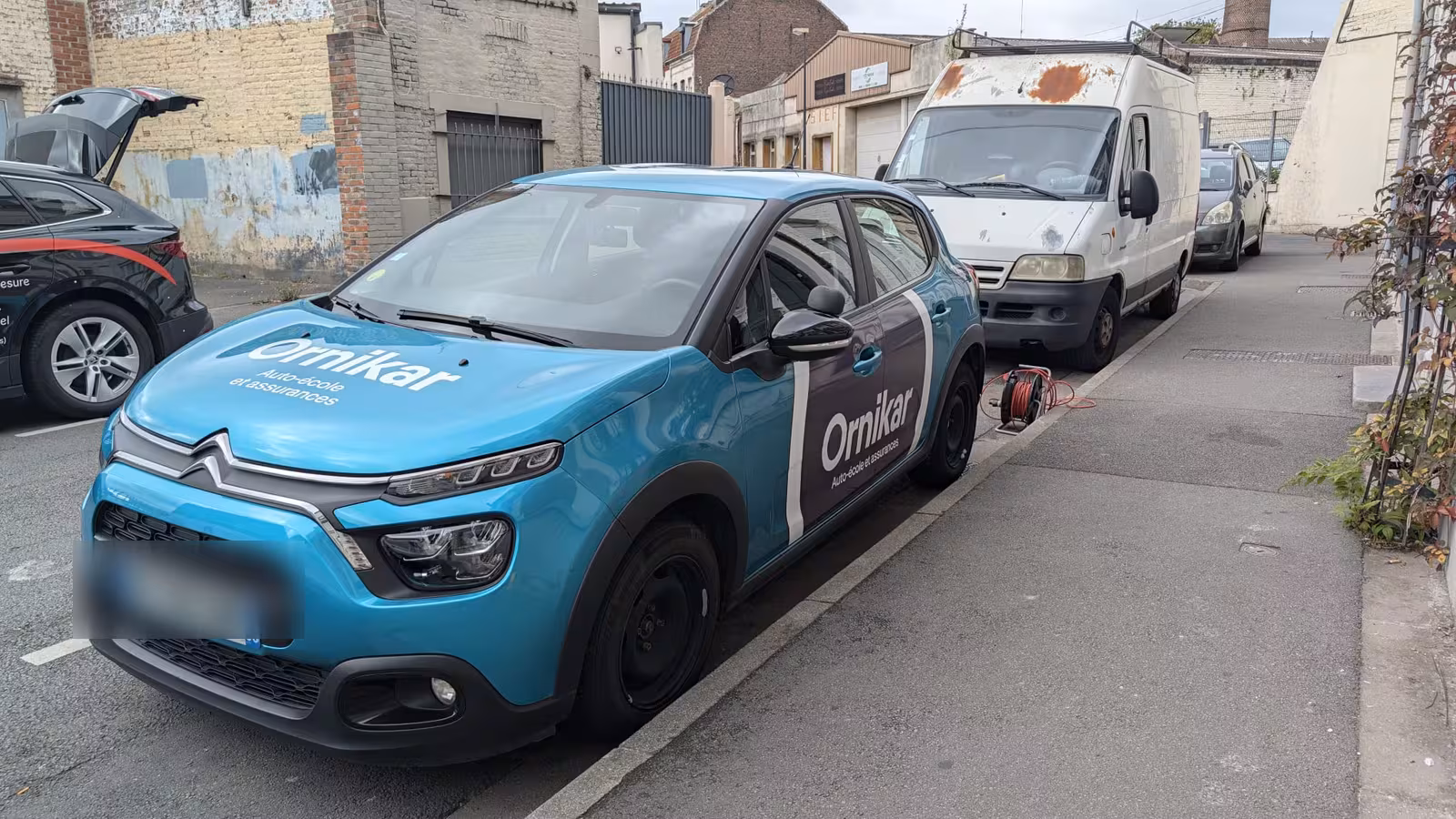 Citroën C3 turquoise et sombre habillée pour la même enseigne, stationnée en rue.